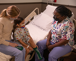 Girl and woman sitting on hospital bed. Healthcare provider sitting next to bed helping girl put on non-skid hospital socks.